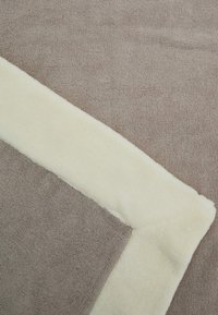 Blød fleece-tæppe med en lys beige kant og en tekstureret brun midte. Kanterne er pænt syet, hvilket øger holdbarheden.