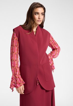 Manteau sans manches bordeaux avec de larges revers et deux poches avant, associé à une blouse rose à manches longues avec un motif et des poignets à volants.