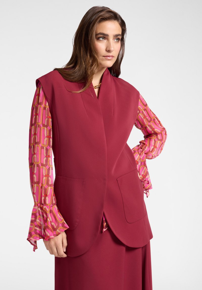 Cappotto senza maniche color porpora con ampi revers e due tasche frontali, abbinato a una camicetta lunga a maniche rosa con motivo e polsini arricciati.