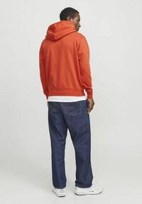 Sudadera con capucha naranja de corte relajado y puños acanalados, combinada con jeans de mezclilla azul oscuro y zapatillas blancas. Diseño simple y funcional.