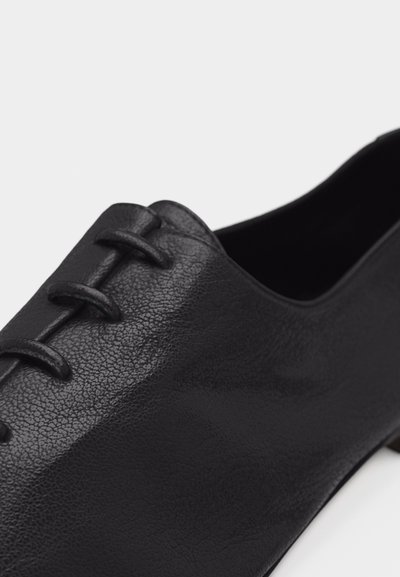 Μαύρο δερμάτινο παπούτσι με λουριά αντικαθιστώντας τα παραδοσιακά κορδόνια, δείχνοντας στενή λήψη της υφής του υλικού και του μπροστινού πάνω μέρους.