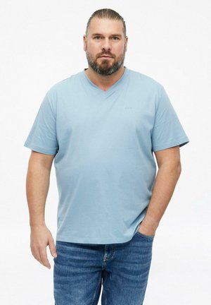 Bearded man with slicked-back hair wearing a light blue short-sleeve shirt and blue jeans, standing with one hand in pocket.