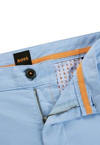 Gai zili bikses ar melnu BOSS etiķeti, oranžiem akcentiem un metāla rāvējslēdzēju. Iekšpusē ir rakstaina oderes apdare bēšā un oranžā krāsā.