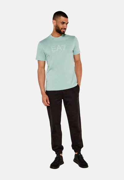 Camiseta de manga corta verde menta con el logotipo en relieve "EA7 Emporio Armani", combinada con pantalones jogger negros y zapatillas negras.