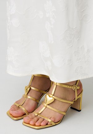 Sandalias de tacón alto doradas con tiras, puntera cuadrada, acentos en forma de corazón y correas ajustables en el tobillo. Llevadas con un vestido blanco bordado.