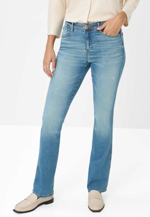 Frau in hellblauen geraden Jeans, beigen Loafers und einem hellen Langarmoberteil, mit einer Hand in der Tasche.