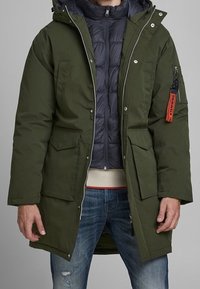 Green parka with a hood, featuring two front pockets, a zip closure, and a black puffer jacket underneath. Notable orange zipper accent.