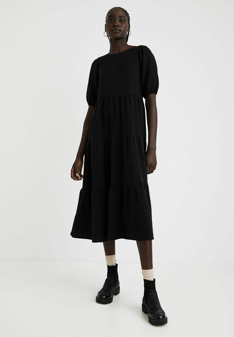 Czarna, warstwowa sukienka z teksturowanej tkaniny z krótkimi bufkami, posiadająca okrągły dekolt i luźną, swobodnie opadającą sylwetkę. Czarne botki za kostkę.