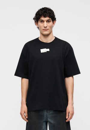 Νέος άνδρας με κοντά σγουρά μαλλιά φοράει υπερμεγέθη μαύρη μπλούζα με μικρή λευκή ετικέτα στο στήθος και σκουρόχρωμο παντελόνι σε απλό φόντο.