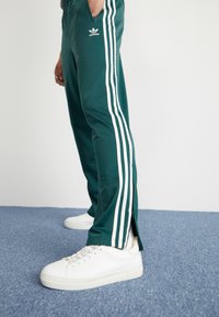 Zelene športne hlače z belimi stranskimi črtami in zadrgo na robovih, ujemajoče se z belimi supergami. Adidasov logotip na zgornjem stegnu. Mehka tekstura.