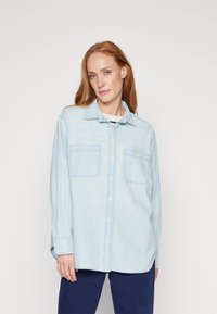 Camicia in denim azzurro chiaro con chiusura a bottoni sul davanti, due tasche sul petto, maniche lunghe e colletto classico. Presenta un tessuto morbido e leggermente strutturato.