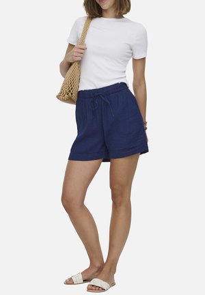 Vrouw draagt een wit T-shirt met korte mouwen, donkerblauwe shorts met trekkoord, witte slidesandalen en houdt een beige geweven schoudertas vast.