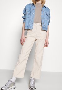 Veste en denim bleu clair sur un t-shirt beige, associée à un pantalon large crème et des baskets argentées. Design simple, coupe décontractée.