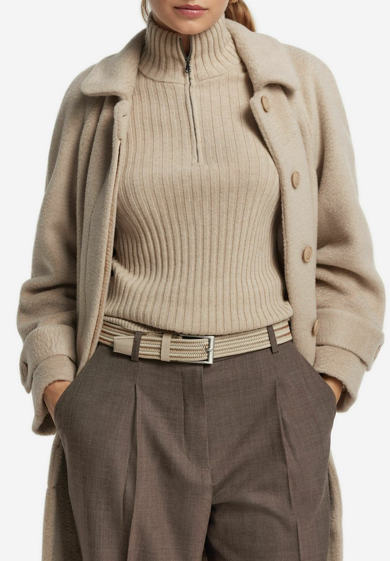 Vrouw in een beige geribbelde ritssweater, bijpassende beige wollen jas met knopen, bruine hooggetailleerde broek en een geweven beige riem. Handen in de zakken.
