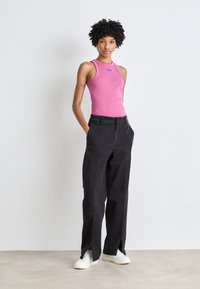Top senza maniche rosa con un piccolo logo, abbinato a pantaloni neri a gamba larga con spacchi laterali. Scarpe bianche completano l'outfit.