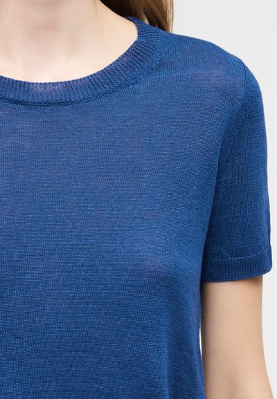 Γυναίκα που φοράει πλεκτή μπλε μπλούζα με κοντά μανίκια και στρογγυλή λαιμόκοψη, εμφανιζόμενη από το λαιμό έως το μέσο του κορμού σε απλό φόντο.