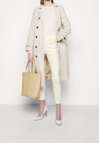 Beige trenchcoat met grote bruine knopen, licht gestreept topje, crème skinny jeans, beige totebag en puntige witte hakken.