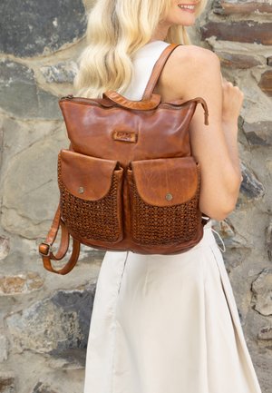 Femme portant une robe blanche porte un sac à dos en cuir marron avec des poches avant tissées devant un mur en pierre.