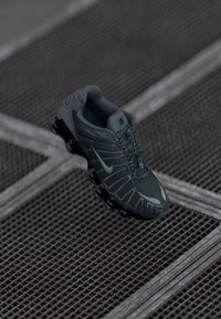 Tamnozeleni Nike tenisice za trčanje s crnim segmentiranim đonom suspendirane iznad metalne mrežne podloge s isprepletenim linijama.