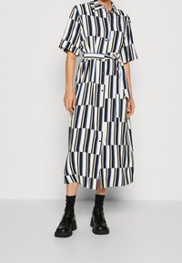 Abito a righe con strisce verticali blu, nere e bianche, maniche corte, chiusura frontale con bottoni e cintura annodata in vita. Indossato con scarpe chunky.