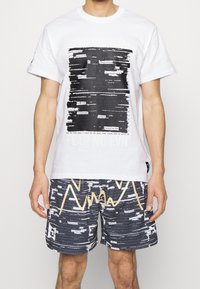 Hvid bomulds t-shirt med sort tekstmønster og "FEAR NO EVIL" tryk. Sammen med sorte og hvide mønstrede shorts med guldaccenter.