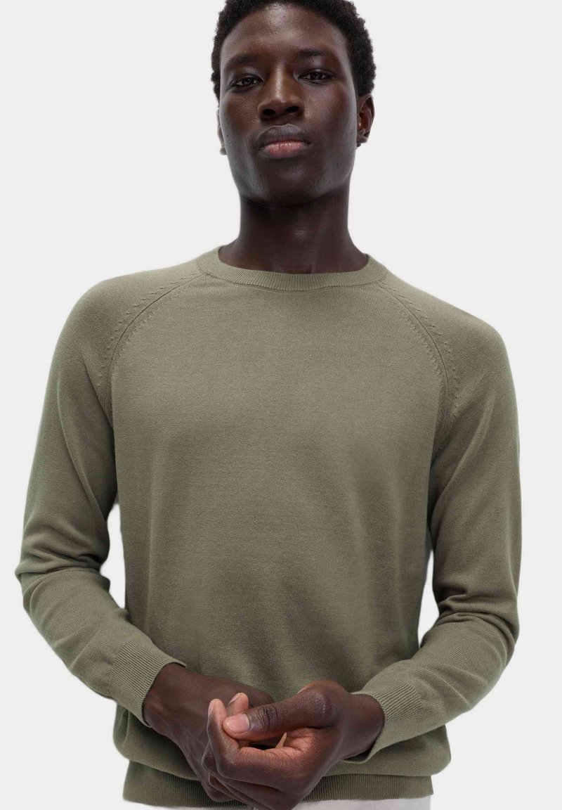 Maglione a maglia verde oliva con scollo rotondo e polsini a coste, caratterizzato da una texture liscia e un design a maniche raglan.