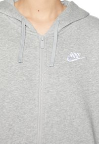 Šedá mikina na zip z měkkého materiálu. Má kapuci se stahovací šňůrkou a malý vyšívaný logo Nike v světle fialové barvě.