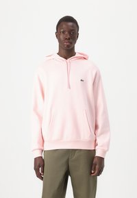 Sudadera de algodón rosa con un bolsillo frontal, capucha con cordón y un pequeño logotipo verde. Combinada con pantalones verde oliva.