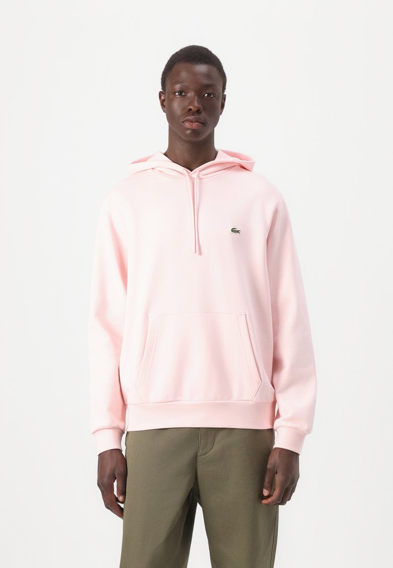 Sudadera de algodón rosa con un bolsillo frontal, capucha con cordón y un pequeño logotipo verde. Combinada con pantalones verde oliva.