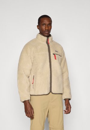 Giacca in pile beige con colletto alto, tasche con zip e dettagli grigi. Presenta una texture morbida e un design aderente.