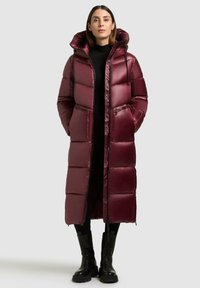 Lang bordeauxrood pufferjack met een glanzende afwerking, grote capuchon, voorkeursrits en twee ritszakken. Draag het over een zwarte outfit en stevige zwarte laarzen.