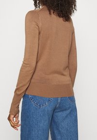 Brązowy sweter dziany z długimi rękawami, prążkowanymi mankietami i dołem, dopasowany krój, noszony z niebieskimi jeansami o wysokim stanie zsercem w kształcie kieszeni z tyłu.