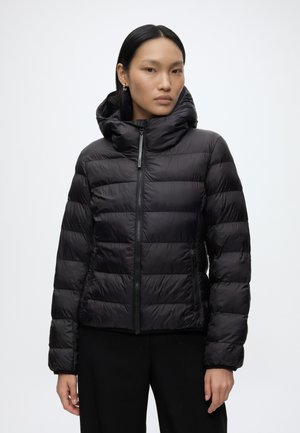 Femme portant une veste matelassée noire avec un col montant et une fermeture éclair, debout devant un fond clair uni.