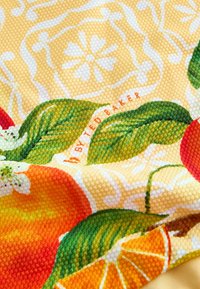 Prim-plan cu țesătură texturată, fructe portocalii, frunze verzi, modele florale albe și textul „By Ted Baker” pe un fundal galben.