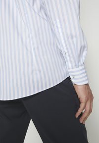 Koszula w jasnoniebiesko-białe paski o gładkiej fakturze, z mankietami na guziki i zaokrąglonym dołem. W zestawieniu z ciemnymi spodniami, widoczna ręka.