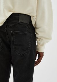 Personne portant un jean en denim noir et un haut à manches longues crème, main détendue le long du corps, sur un fond clair uni.