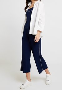 Námořnický pletený dvoudílný outfit, který obsahuje přiléhavou tílku a široké zkrácené kalhoty s bočními rozparky, zakrytý bílou košilí s knoflíky.