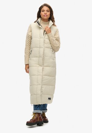 Beige langer Puffer-Westen mit hohem Kragen, gesteppter Struktur, Seitentaschen und Markenlabel, getragen über einem cremefarbenen Pullover und blauen Jeans.