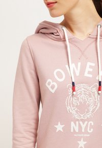 Kvinna iklädd en rosa hoodie med vita dragskon, med ett broderat tigeransikte och texten "BOWERY NYC Hilfiger Denim."