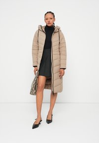 Béžová péřová bunda s kapucí, která má diagonální prošívaný design, přední zip a nastavitelné šňůrky. Spárováno s černým rolákem a sukní.