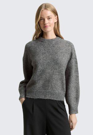 Jeune femme aux longs cheveux blonds portant un pull en tricot gris et un pantalon noir, debout avec une main dans la poche devant un fond uni.