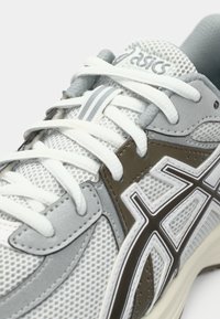 Zapatilla deportiva blanca y gris con material de malla, forma redondeada, suela de goma, acentos marrones en contraste y un sistema de cordones destacado.