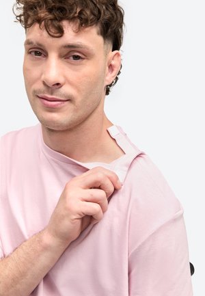 Muškarac kovrčave kose u ružičastoj košulji, povlači vratni izrez unatrag kako bi otkrio bijelu etiketu ili kopču na ramenu.