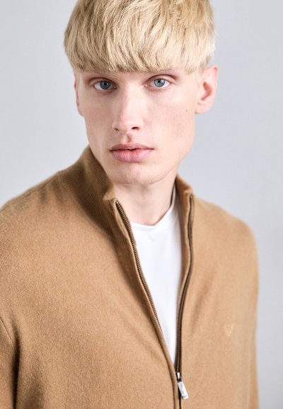 Jauns vīrietis ar blondiem matiem un zilām acīm, ģērbies smilškrāsā apkakles džemperī virs baltas krekla, skatās tieši uz kameru.