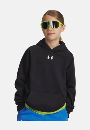 Noor inimene, kes kannab neoonrohelisi peegeldavaid päikeseprille, musta Under Armouri kapuutsiga dressipluusi ja siniseid lühikesi pükse, seisab käed taskutes.