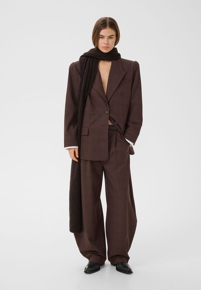 Jeune personne portant un costume marron oversize, écharpe noire enroulée autour du cou, debout avec une main dans la poche sur un fond uni.