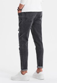 Hombre con pantalones vaqueros rasgados de color gris oscuro y zapatillas blancas, de pie con las manos en los bolsillos frente a un fondo liso.