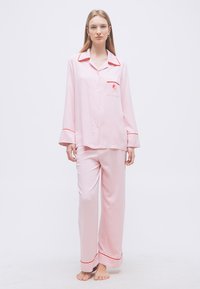 Νεαρή γυναίκα στέκεται ξυπόλυτη φορεμένη με ροζ μετάξινες πυτζάμες με κόκκινη επένδυση και μικρή τσέπη στο στήθος.