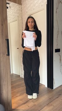 Young woman in black top and pants stands indoors holding folded white fabric against a textured wall and wooden floor background.