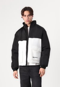 Black and white insulated jacket with a high collar, front zipper, and two large pockets. Features elastic cuffs and a logo on the hem.